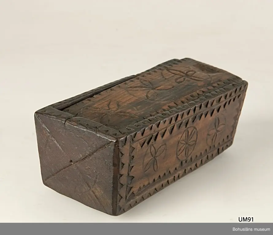 Rektangulär låda med skjutlock och något snedställda kortsidor.
Skuren dekor bestående av rosetter, kors samt uddsnitt längs kanterna.
På en av långsidorna är årtalet "1848" och initialerna "AN" inskurna.
Inuti ligger en äldre etikett från ca sekelskiftet.
Kan vara en friargåva.

Litteratur:
Gjaerder, Per, Esker og tiner, C. Huitfeldts förlag, Oslo, 1981, s. 41.
Knutsson, Johan, Friargåvor, Nordiska museets förlag, Stockholm, 1996.

Ur handskrivna katalogen 1957-1958:
"Trälåda
1870-1887
Bottenmått: 14 x 5,5 H. 5. Trä med skuren ornering. Föremålet helt. Bohusl."

Lappkatalog: 84