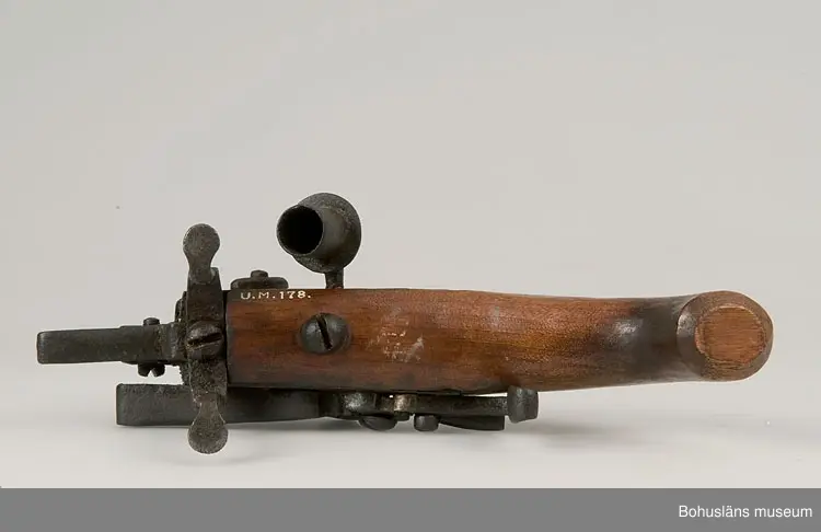 Ur handskrivna katalogen 1957-1958:
Elddon, "1800".
L. 22, H. c:a 13 cm; ett flintlås monterat m. handtag av trä och 2 fötter av järn; på sidan en lucka på gångjärn, m. regel, på denna (d.v.s, på luckan) en ljushållare av järn. Helt.

Lappkatalog: 90