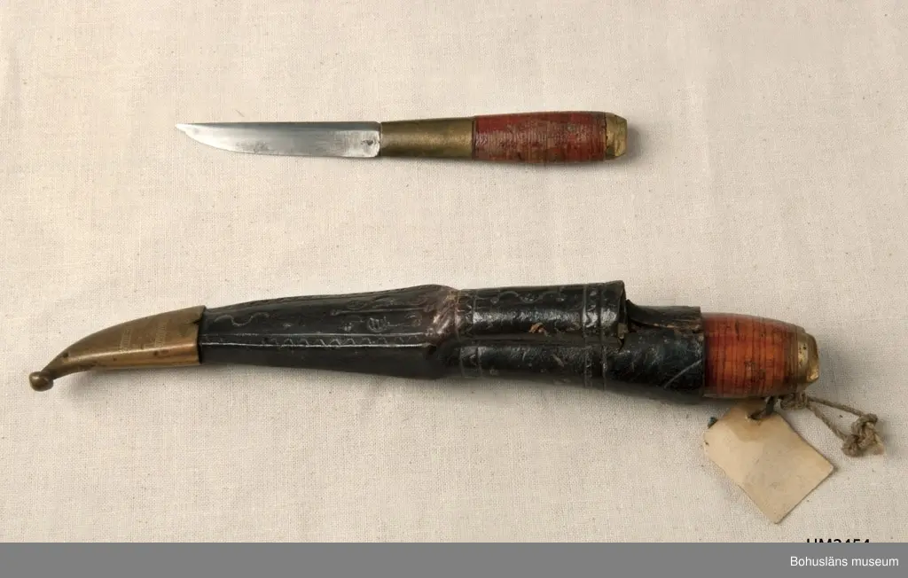 Två knivar i en och samma slida. En större (längd 16 cm) och en mindre (längd 12 cm).
Knivarnas skaft är gjorda av näver som packats tätt kring tången.
Lädret i slidan har pressat mönster och har färgats svart. Mässingstoppen har ristad/graverad dekor. Slidan är fodrad med ett ljust gröntonat papper.

Knivarna är tillverkade i Kalajoki, en stad i Norra Österbotten i Finland. Slidans doppsko är tillverkad i en s.k. Kauhava-modell. Kauhava är en stad i Södra Österbotten .

Ur handskrivna katalogen 1957-1958:
Två slidknivar i en slida
Kniven a) L. 16 cm; m. skaft av mässing och läder; kniven b) L. 11,8 cm; lik föreg. Slidan c) L. 16 cm; svart läder, skoning av mäss. Föremålen hela.

Lappkatalog: 29