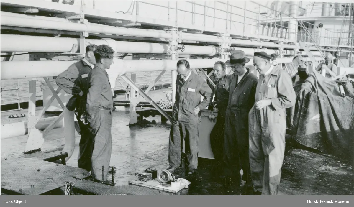 Menn som studerer en slags innretning på dekk på tankskipet M/S Haakon Hauan, B/N 465 9. juli 1935. Mannen ytterst til høyre er Hr. R. Käsler (?), og mannen nærmest innretningen er Hr. Steinhauser (?). Skipet ble levert av Akers Mek. Verksted i 1935 til Østlandske og Vestlandske Petr. Co.