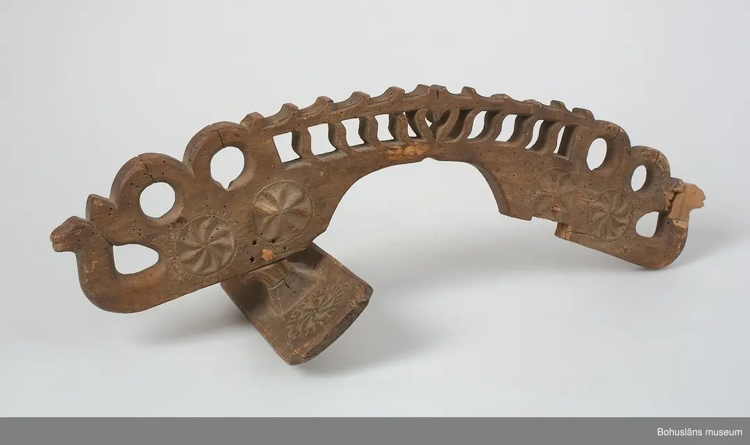 Böjd, genombruten båge med karvsnittsdekor på båda sidor bestående av rosetter. Längst ut på vardera ända är ett drakhuvud.
Rektangulära fästen, vilka vilar på hästens manke. Karvsnittsdekor med rosetter på en av dem. Den andra är sekundär och har skurna cirklar samt är defekt.
Ett drakhuvud är sekundärt.
Skadedjursangrepp.

Litteratur: Edvardsson, Ingmar, Rack, ryss och rissla, Stockholm, 1995, 
s. 66 samt bild s. 42.
Kjellberg, Sven T.: Svenska selkrokar av trä i Nordiska museet. Fataburen 1926. 
Nylén, Anna-Maja, Hemslöjd, Håkan Ohlssons förlag, Lund, 1978, s. 396-401.

Ur handskrivna katalogen 1957-1958:
Selranke av trä
I 3 delar a-c, a) = oket L. 59 cm; b) "stödplatta", L. 19,5 cm; c) d:o, L. 20 cm (nygjord). Oket har snidad dekor i genombrutet arbete m. drakhuvud i ändarna (ett nygjort); hela hela selranken har karvsnittsdekor. Omålad. Maskäten. 

Lappkatalog: 40
Hugo Hallgren var son till salteriidkaren och handelsmannen Johan Jacob Hallgren och Emelia Gerle. Företaget började sälja brissling på 1850-talet och 1864 startade man konservfabrik på Gullholmen som senare döptes till J. J. Hallgren & Söner. Sönerna var Gustav Richard, f. 1858 och Oscar Hugo, f. 1867.  Här introducerades flera nyheter inom ansjovisindustrin,  bl a Sveriges första skinn- och benfria delikatessill. Företaget var också först med att lägga in ansjovis i hermetiskt slutna kärl av bleckplåt. 1898 köptes Hallgrens konservfabrik upp av det nybildade storföretaget AB Sveriges Förenade Konservfabriker (SFK). 1901 flyttade Hugo Hallgren till Ellös där han startade en ny konservfabrik.



För uppgifter om förvärvet, se UM5155.