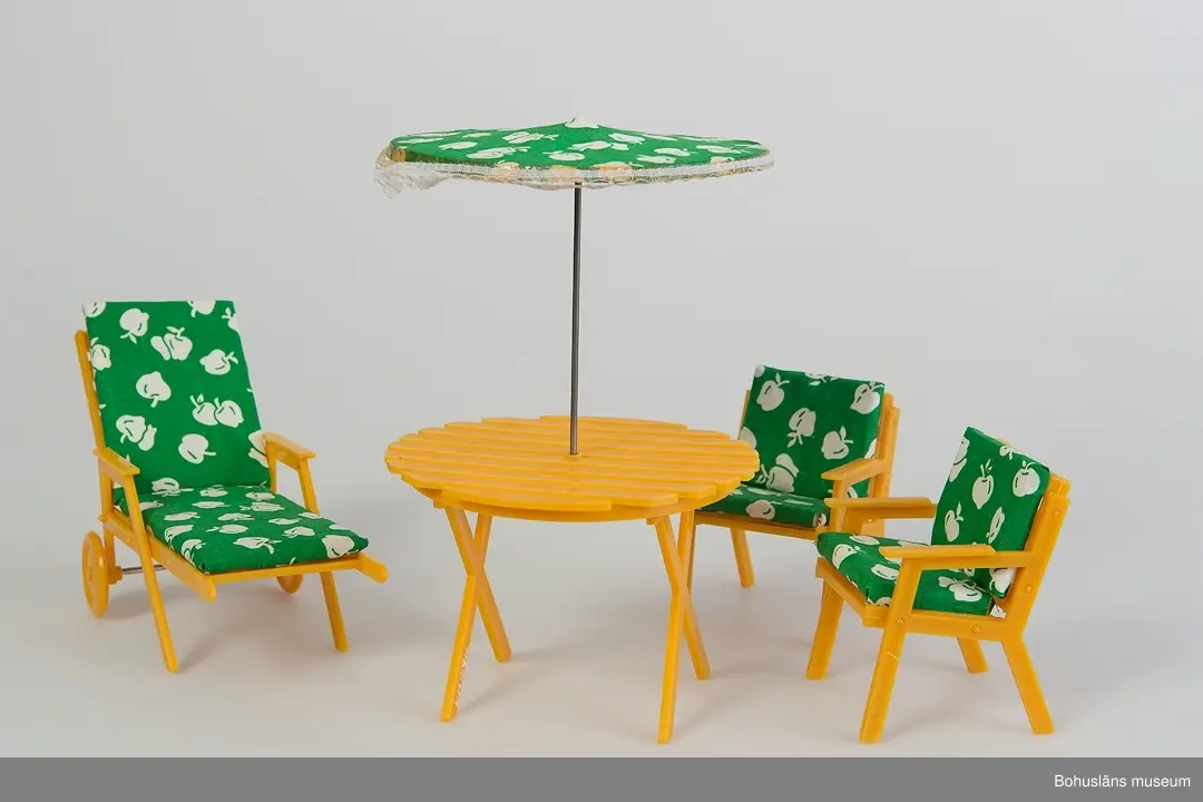 Trädgårdsmöbel bestående av bord med parasoll, två karmstolar samt en vilstol med ett par hjul. Alla objekten i flera delar som sammanfogas.
Dynorna till stolarna är i ett mönstrat grön-vitt tyg. Parasollet har samma tyg som dynorna med kantdekor av spets påsatt. Totalt antal delar: 25 st.
Metallpinne till parasoll saknas. Den som ligger tillsammans med möbelns delar är rekvisita.

Se UM015810