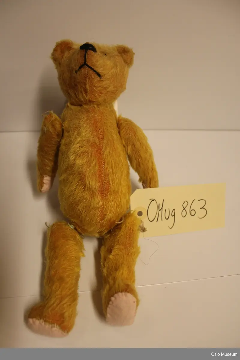 Mellomstor lys oransje bamse i halm, trukket med kunstig "pels", alle lender er bevegelige. Knapper som øyne, påsydd snute og munn.