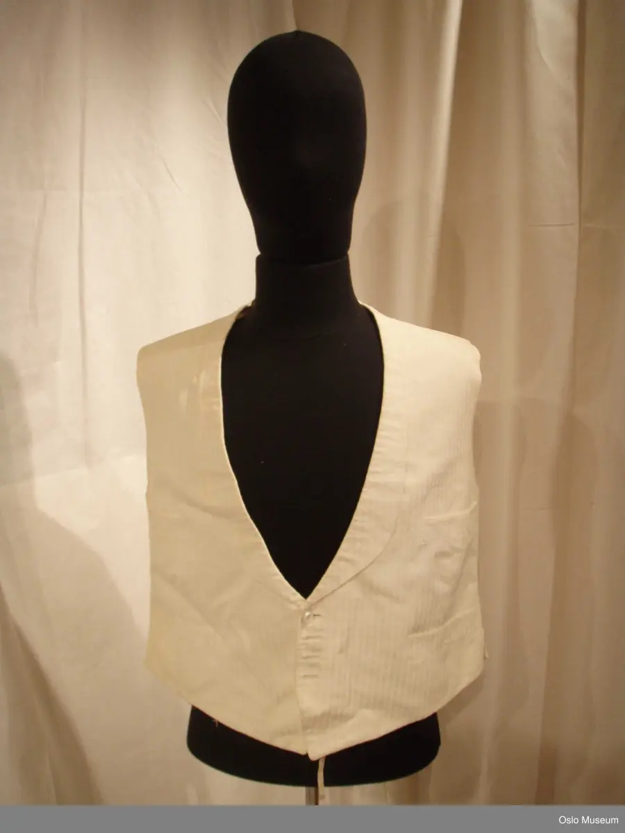 1: Vest i hvitt stoff med innvevde striper i brystparti. Knepping i form av 1 hvit knapp i perlemor, 3 mangler. Spenne bak.
2: Vest i hvitt stoff med innvevde ranker/blomster. Knepping foran med 3 hvite runde perlemor (?) - spenne bak