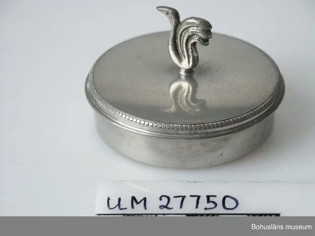 Föremålet visas i basutställningen Uddevalla genom tiderna, Bohusläns museum, Uddevalla.

Rund dosa med plant lock med nedvikt kant. På dosans lock en knopp i form av en stiliserad svan. Lockets ytterkant dekorerat med ytterst små rundlar i upphöjd relief. Stämplad undertill: " SVENSKT TENN / L med två kronor, / U / E8". (E8 = 1931)

Dosan representerar ett stycke hantverkshistoria från Uddevalla. Högklassiga produkter spreds över hela Sverige från juvelerarfirman Harald Linder. Guldsmed Linder drev dessutom firman AB Svensk Metallkonst grundad ca 1911 - 1912, guldsmedsaffär, present- och prydnadsaffär och tidvis också "konditorirestaurang" i Uddevalla. Han hade också två tennbutiker, en i Stockholm öppnad 1930 och en i Göteborg, startad 1928. Ca 40 - 50 personer arbetade i Uddevalla med att tillverka tennföremål för Linders båda affärer "Tenn Lådan" i Göteborg öppnad 1928 och i Stockholm öppnad 1930.
För en mer utförlig beskrivning av Harald Linder, se dokumentation i Bilagepärmen UM26919.

Dosan är en testamentarisk gåva av Birgit Björkholm, född och uppvuxen i Uddevalla och vars far Oscar Björkholm drev urmakeri i staden, se UM64.05.001 - UM64.05.119, och  Arkivnummer 172.

Föremålet kompletterar intervjumaterial från projektet "Arbetarstaden
Uddevalla" 1998. Se museets arkiv; Etnologiska undersökningar, seriesignum M 11 M.