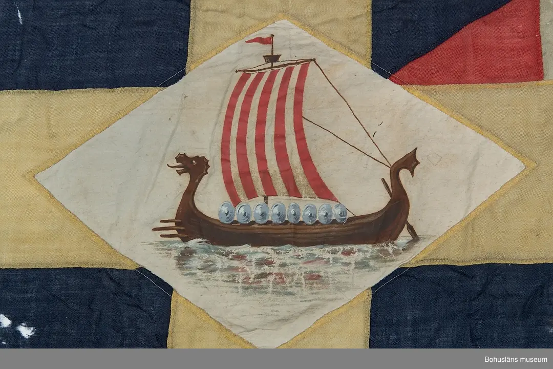 Segelsällskapet Vikens flagga, SS Viken. 
Svensk tvåtungad  flagga med parti "Silllsallaten", svensknorska unionsflaggan i övre hörnet samt infällt handmålat rombiskt emblem placerat mitt i det gula korset. På emblemet är målat ett vikingaskepp. 
Märkt "KASEN" med svart märkbläck. Hand- och maskinsömnad.
Skador. Eventuellt har flaggans form ändrats.