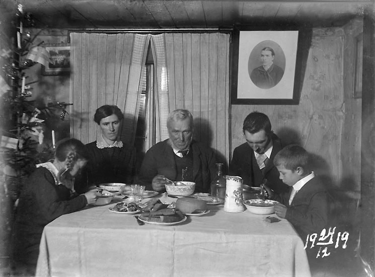 Familjen på Skistad gård äter julmiddag. Runt bordet sitter från vänster Lisa, Klara, Johannes A, Johan Johansson och Gösta. Fotografiet på väggen föreställer Christina Johansdotter, Johannes avlidna hustru. Johan och Klara är barn till Johannes och Christina, Lisa och Gösta är fosterbarn till Klara och Johan.