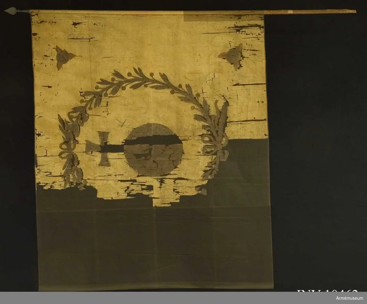 Fanduk av vitgul (röd) taft med målade emblem: Upplands sköldemärke: ett riksäpple i brunt, svart och gult, allt inom en lagerkrans av två kvistar med tätt ställda blad, nedtill hopknutna med bandrosett. I hörnen brinnande fyrkulor i svart och rött. Ursprungligen fastspikad med sidenband och kupiga mässingstift, nu fastspikad med några spikar och f ö fastsydd. Stång av brunpatinerad furu. Spets av järn, troligen sekundär, holken enkel, bladet massivt.