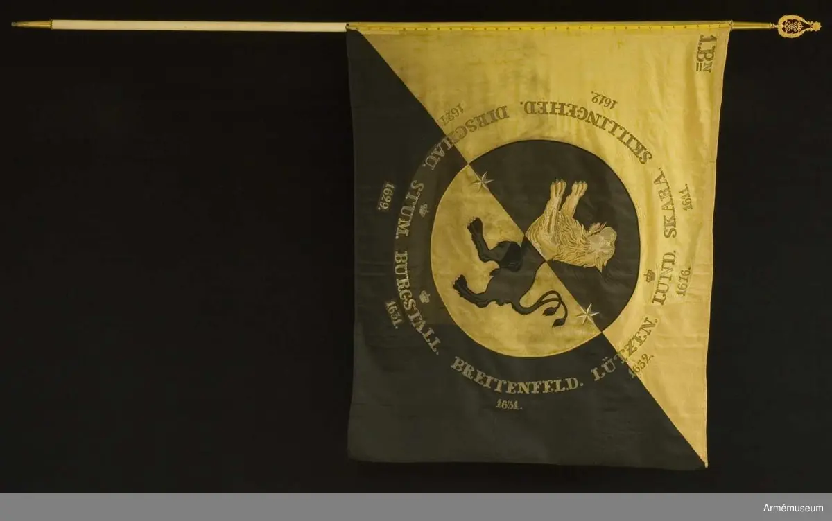 Duk av fansiden i fyrskaftad kypert, delad diagonalt i två fält - det inre gult och det yttra svart. I mitten en cirkel med 800 mm radie med omvända färger och där inne Västergötlands sköldemärke: ett lejon, gult på det svarta och svart på det gula fältet. I fältens delning finns två sex-uddiga stjärnor i guld. Lejonets svarta är applicerat fansiden  med kantsöm i svart silke, det gula är dels applicerat fansiden, dels schattérsöm i gult och vitt siden. Tungan fodrad i rött silke. Omkring vapnet, genombroderat i guld, finns segernamn: SKARA med "1611" under, SKILLINGEHED med 1612, DIRSCHAU och1627,  krönt STUM och 1629, krönt BURGSTALL och 1631, BREITENFELD och  1631, LÜTZEN och 1632 samt krönt LUND med 1676. I övre inre hörnet finns "1.Bn" broderat i guld (två streck  under N). Duken är kantad med ripsband, svart på det svarta och gult på  det gula. Fastspikad med förgyllda spikar. Kravatt av gult och svart kypertvävt fansiden. Guldfrans på kortändarna.

Hel längd 2860 mm, frans 10 mm, bredd 260 mm. Stång av furu - vitmålad. Doppsko konisk med rund knopp av förgylld mässing. Längd 2560 mm. Spets av förgylld mässing, ursprungligen med Oscar I:s krönta monogram som senare felaktigt byttes mot Gustav V:s krönta spegelmonogram inom lyra av lager. Längd 330 mm. Fodral av mörkblått (svart) kläde med skinnskodd topp, dragsko och ficka för spetsblad. Längd 1820 mm, bredd 180 mm.