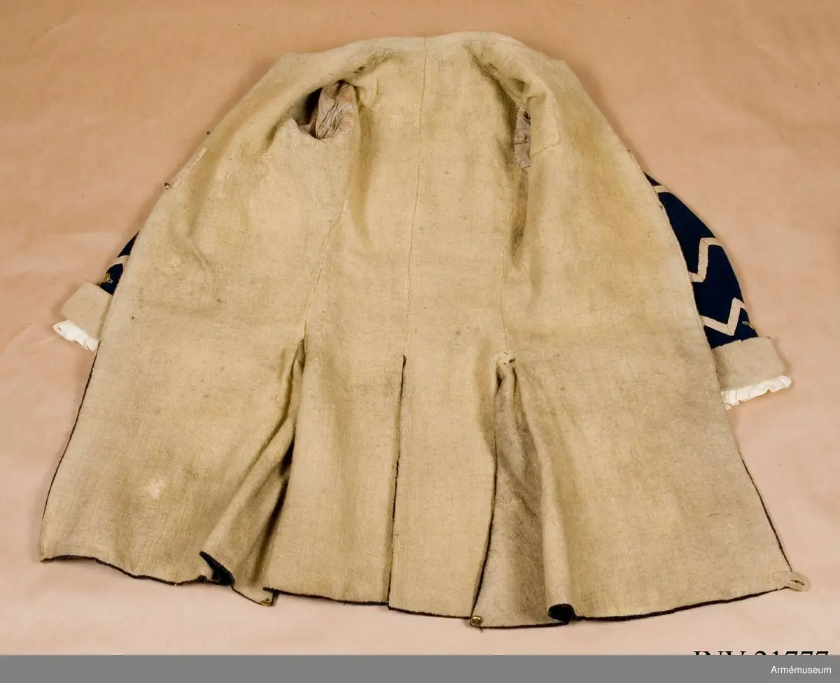 Rock av blått kläde med vita ärmuppslag och bröstrevärer av vitt kläde, gula axelkarmar, dekorationer av vävda vita band i tre olika bredder och släta mässingsknappar. Rocken är lång, med svängd mitt-fram-linje. Troligen nådde den nästan till knät på den som bar den. Den har 20 parvis ställda knappar fram, men knäpps kant i kant med två hyskor och hakar.
Den är konstruerad med hela framstycken som går runt till baksidan och bildar sidstycken. Delat bakstycke med mitt-bak-söm, samt tvåsömsärm. Sidsömmarna, ärmsömmarna och mitt-bak-sömmen är dekorerade med band. Två lågt ställda horisontella fickor fram med bandprydda ficklock, falska knapphål och två knappar vardera.
Skört med uppvik nertill fram. Sicksackdekorationer och rundlar på ärmarna.
Foder av vit boj i bålen, men oblekt bomull i ärmarna.