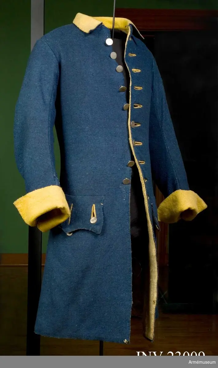 Rock av blått kläde med gul krage och ärmuppslag, fodrad med gul boj i livet samt linne i ärmarna. Besatt med tennknappar. Märke efter lacksigill.