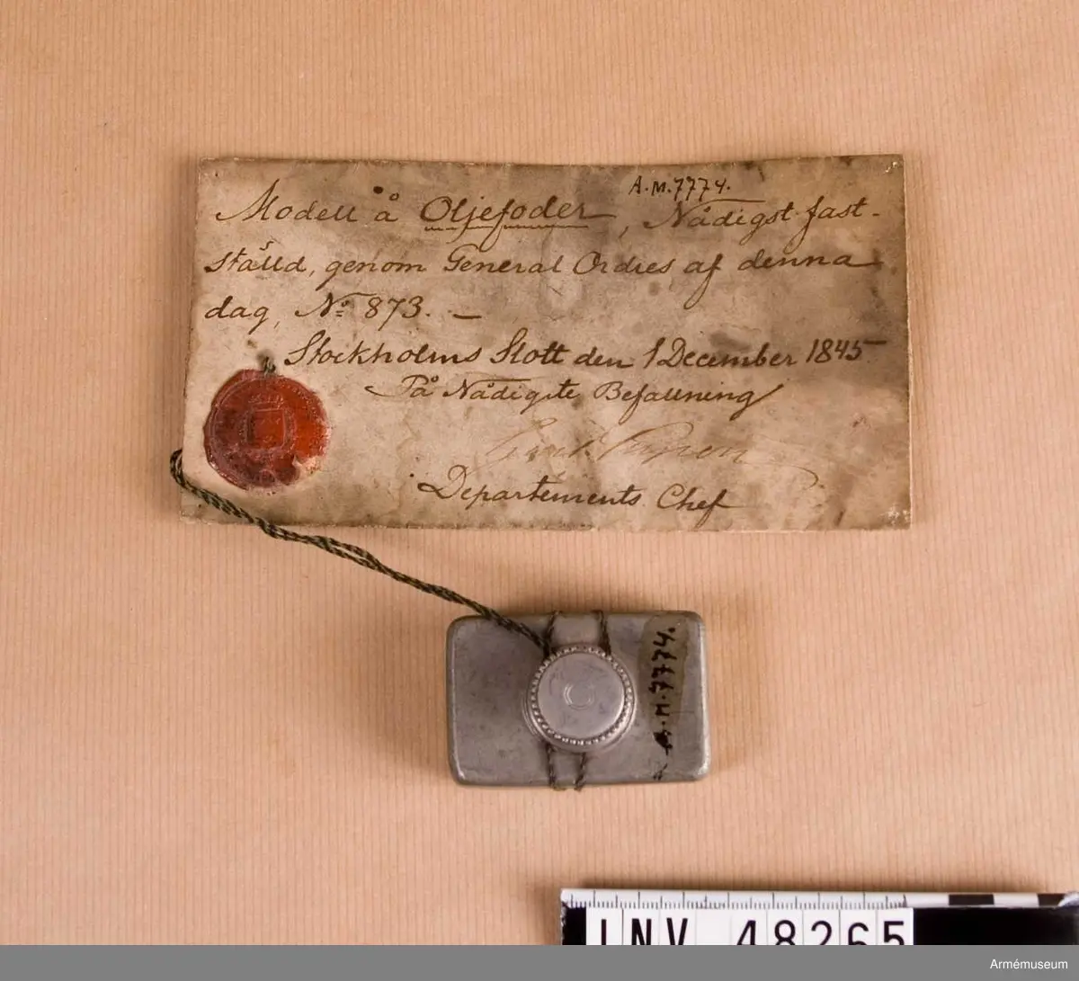 Fyrkantigt oljefoder av huvudmodell.
Text på modellappen:
"Modell å oljefoder, Nådigst fast-
ställd, genom General Ordres af denna 
dag, No 873.-
Stockholms Slott den 1 December 1845
På Nådigste Befallning
   Carl ......?
Departements Chef"