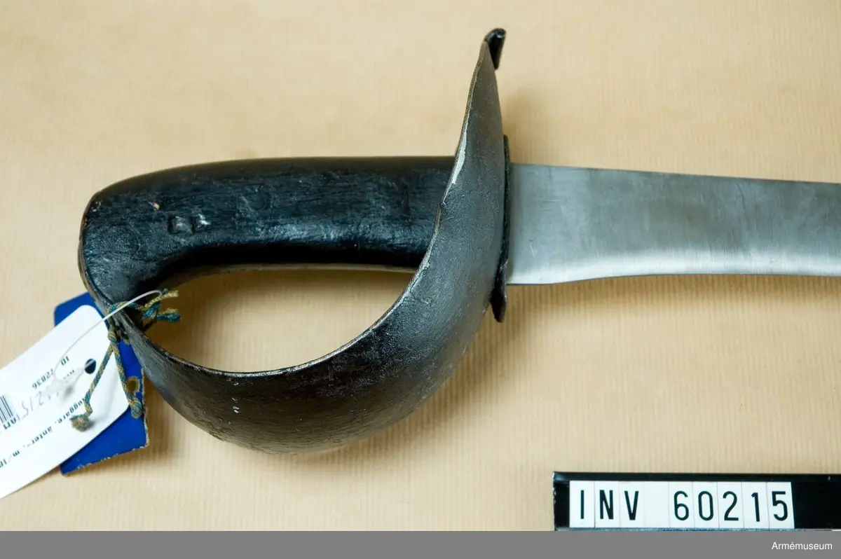 Grupp D II.  
Klingan är av samma typ som på AM 60213 och av Hafströms  konstruktion. Upptill är den 43 mm bred, men 120 mm längre ned  är bredden blott 33 mm. Sedan tilltar den så småningom, så att  450 mm, längre ned är den 42 mm, varefter den åter avtar mot  udden. 250 mm ovanför udden får även ryggsidan egg, som  fortsätter till udden.Tången är bred och upptill framåtböjd. Kaveln bildas av ett par svartmålade träskenor, vilkas hålls fast vid tången genom tre nitar. Parerplåten är av svärtat järn, rätt stor, något uppböjd samt på yttersidan betydligt större än på innersidan. Baktill  utgår från parerstången en kort, rektangulär, baktill nedböjd  och med förstärkt bakre kant försedd flik. Framåt blir  parerplåten smalare samt tjänstgör som handbygel.Längst upp ligger dess övre ände ovanpå kavelns övre ände och fasthålls där genom att tången, som går upp genom ett hål på handbygeln är nitad på översidan