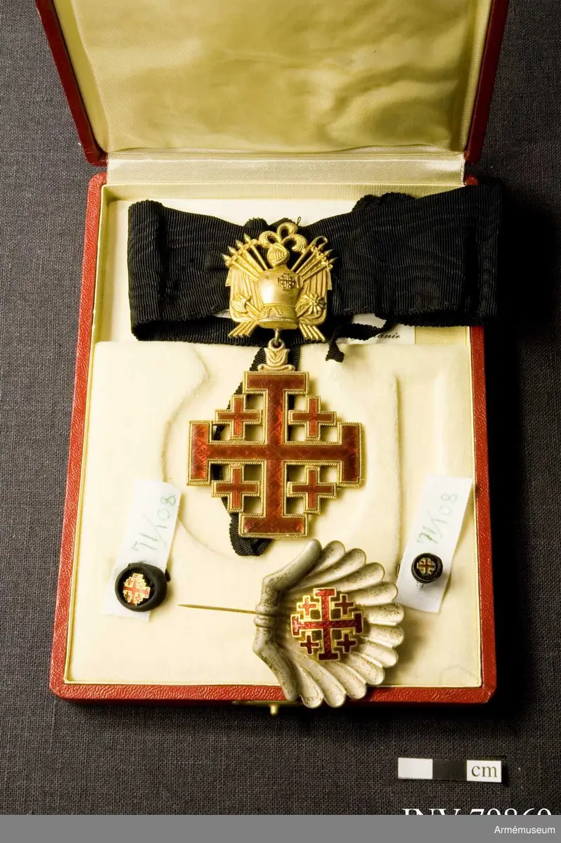 Påvliga Heliga Gravens orden, kommendörstecken i svart band. 
Miniatyr och knapp. Ordenstecknet i form av pilgrimssnäcka tilldelades ordensriddare som genomfört en pilgrimsresa till det heliga landet.

Förvaras i samhörande etui.