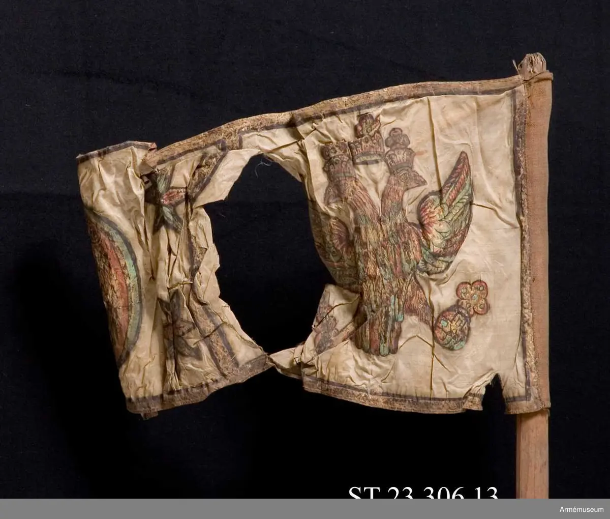 Duk av vit sidentaft och strumpa av rött linne. Motivet målat i guld- och silver samt rött och grönt bestående av en krönt örn, måne, sol och stjärnor, På duken en pappersetikett som anger att den tagits i slaget vid Saladen 1703.