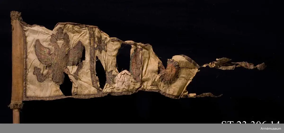 Duk av vit sidentaft och strumpa av rött linne försedd med frans vid öppningarna. Motivet målat i guld- och silver samt rött och grönt bestående av en krönt örn, måne, sol och stjärnor, På duken en pappersetikett som anger att den tagits i slaget vid Saladen 1703.