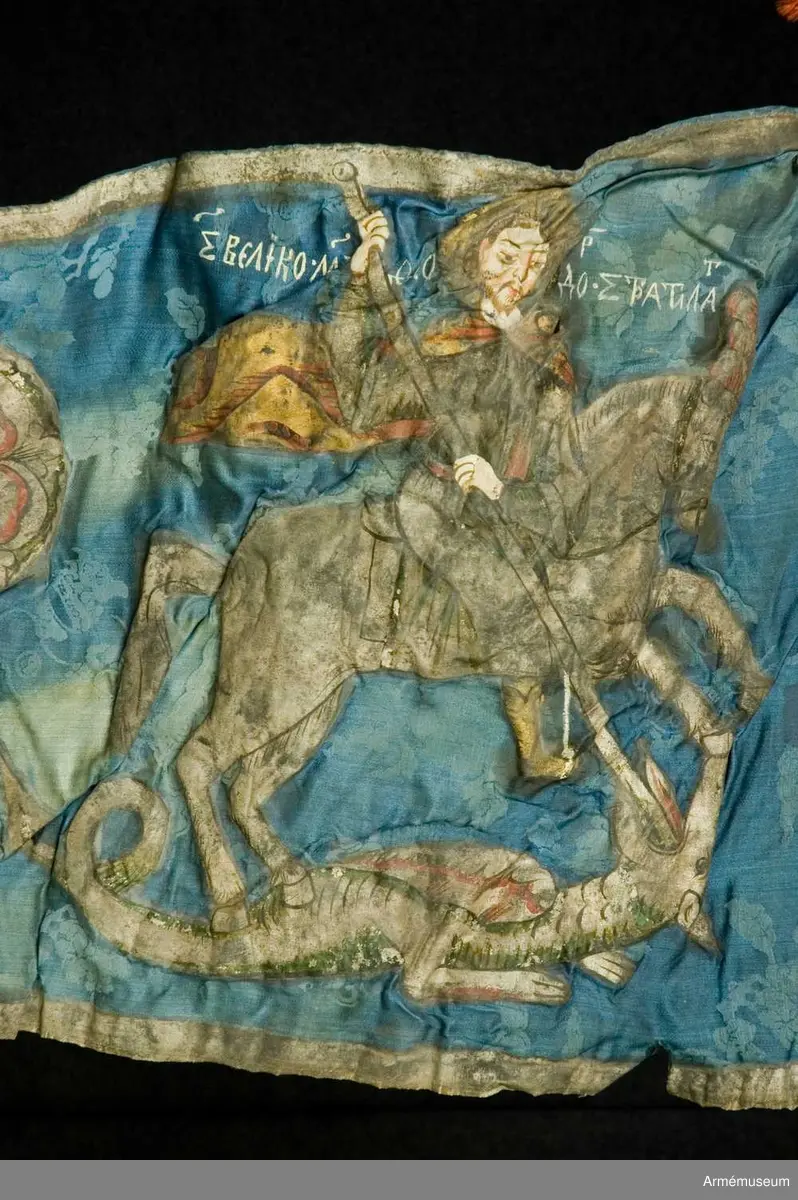 Entungad lansfana. Duk av blå kinesisk sidendamast med målad dekor. I 1703 års inventarium anges motivet vara St Göran men enligt den ryska inskriptionen är det Feodor Stratilat . Rosor och stjärnor av silver. Duken kantad med en silverbård. Strumpa av rött linne.