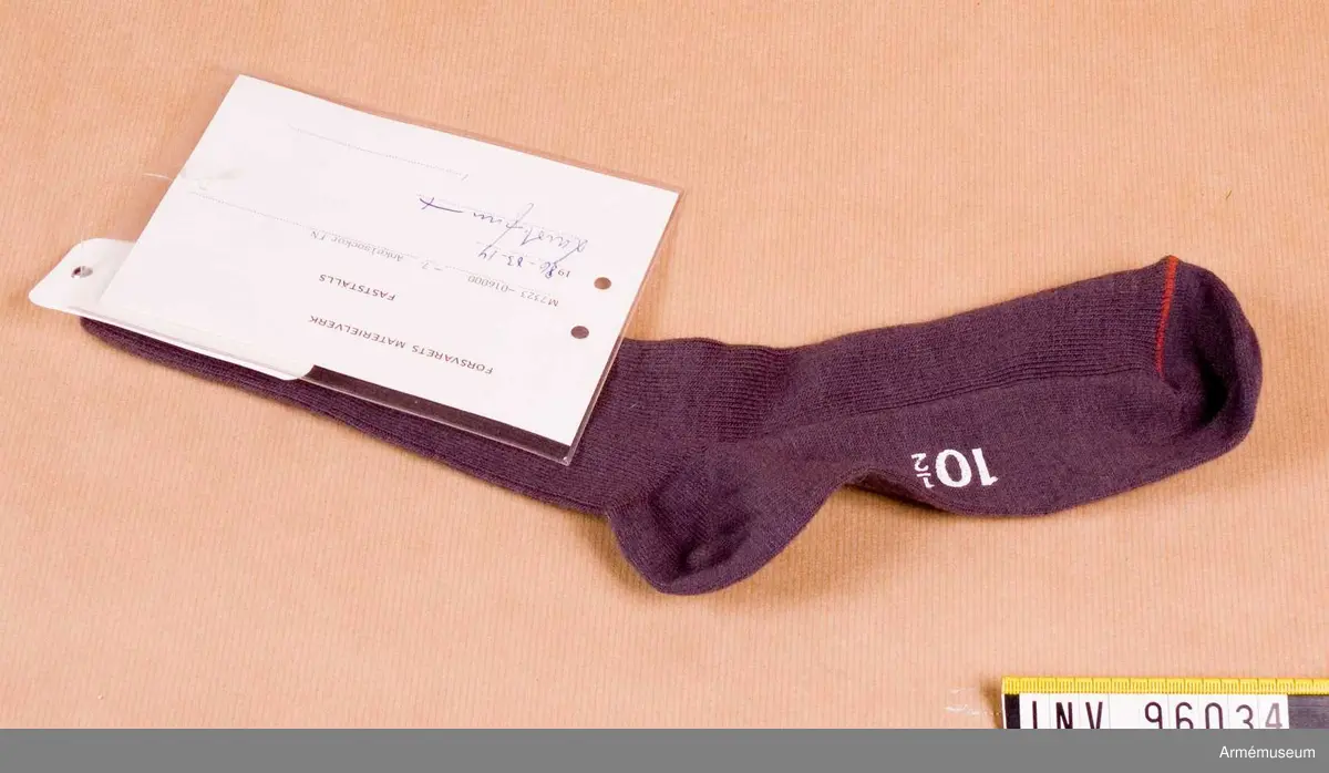 Vidhängande etikett med text: "Försvarets Materielverk, Fastställs, M 7323-016000-7, Ankelsockor FN, 1986-03-14, (oläslig underskrift)"