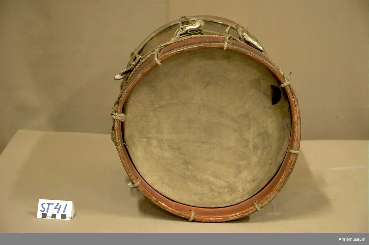 Cylindertrumma av : ljus metall, dekorerad. Metallen är bucklig och har ett mindre hål.

Membran : båda skinnen är trasiga. Ovanpå det övre skinnet ligger en färgad pappskiva. Det undre skinnet har en skada, ca 4 cm. med en flik som går inåt.

Veckelgjordar : träet skiktar sig i den övre veckelgjorden.
 
Spännlina : ser hel ut med 8 st. spännstroppar av vitfärgade tjocka  läderband ca 3 cm. breda. Tre av dessa har tandklippta avslut. 

Spännringar : finns.

Bärring : 

Utsmyckning : vapensköld med mansfigurer på vardera sidan.

Märkning : finns.