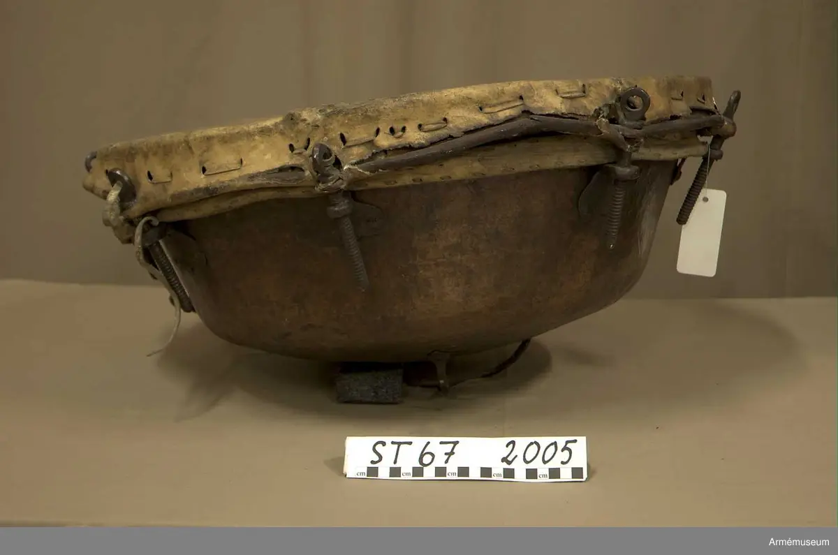 Kittel av koppar och skinn av kalv med fast spännring. Fot av järn fastnitad med kopparnit. Skruvar och skruvfästen av järn. Bärrem av kalvskinn i två lager med spänne av järn.