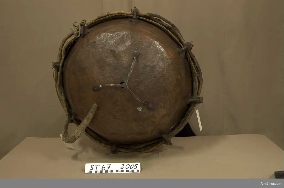 Kittel av koppar och skinn av kalv med fast spännring. Fot av järn fastnitad med kopparnit. Skruvar och skruvfästen av järn. Bärrem av kalvskinn i två lager med spänne av järn.