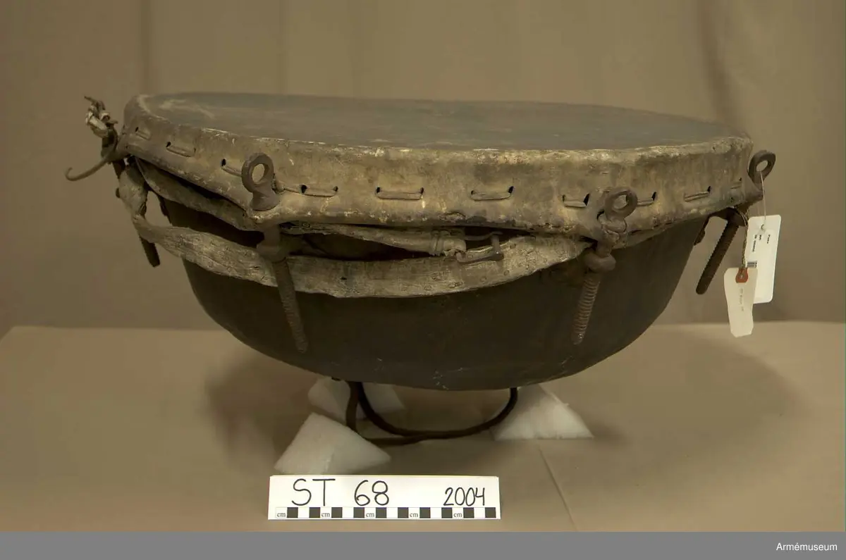 Kittel av koppar och skinn av kalv med fast spännring. Tredelad fot av järn. 7 skruvar och skruvfästen av järn. Bärrem av kalvskinn i två lager med spänne av järn.