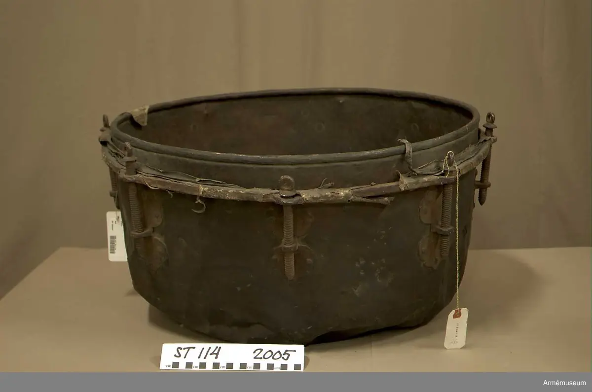 Tillstånd 2005:
Kittel: kraftigt bucklig och sprucken på flera ställen. En mängd stickmärken i storlek 1 cm. - 4 cm. Fot: saknas. Spännskinn: saknas, förutom en liten flik - hängandes över kanten in i pukan - likaså är skinnet lämnat längsmed kittelkanten fastsytt genom ett smalt läderband. Spännring: rostig. Spännskruv: 8 st. Skruvfästen: 8 st. Bärrem: saknas.