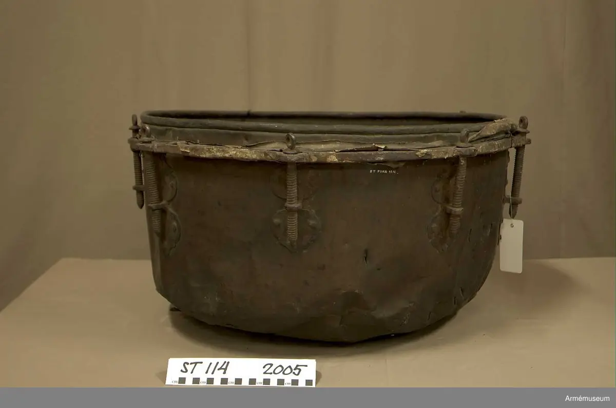 Tillstånd 2005:
Kittel: kraftigt bucklig och sprucken på flera ställen. En mängd stickmärken i storlek 1 cm. - 4 cm. Fot: saknas. Spännskinn: saknas, förutom en liten flik - hängandes över kanten in i pukan - likaså är skinnet lämnat längsmed kittelkanten fastsytt genom ett smalt läderband. Spännring: rostig. Spännskruv: 8 st. Skruvfästen: 8 st. Bärrem: saknas.