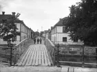 Tre män poserande vid gångbron över ån vid Munksundsgatan, Enköping, vy från nordöst och Hamngatan, 1907.