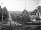 Rosenborgsgatan i Huskvarna år 1910. Tvåvåningsvillor i trä ligger utmed den trädkantade vägen och häckar inramar trädgårdarna. I förgrunden syns järnvägsspår.