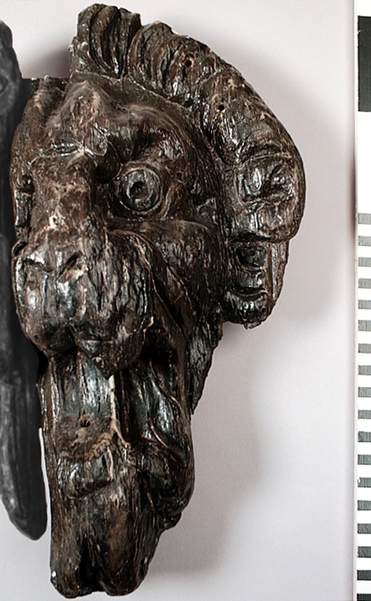 Skulpturdel, höger sida av lejonmaskaron. Lejonmaskaronen är i sin helhet rektangulär till formen, och utförd i två, eventuellt tre, sektioner. Lejonet har stora, runda ögon under kraftiga ögonbrynsbågar. Nos- och käkparti är kraftigt, med V-formade näsborrar och ett vidöppet gap som blottar spetsiga tänder och en bred tunga. Den i förhållande till ansiktets storlek relativt obetydliga manen gör ett stubbat intryck med markant vågformig ytterkant. Hårtestarna är återgivna i mjuka slingor i bakvänd S-form.

Lejonmaskaronen är kraftigt sliten, med sprickor. Mindre partier av mankransen saknas.



Text in English: Part of a sculpture, the right side of a lion mask. The lion mask is I SIN HELHET rectangular in form and made in two, perhaps three, sections. The face has large round eyes with heavy eyebrows and a powerful nose and jaws, V-shaped nostrils, wide-open mouth revealing pointed teeth and a wide tongue. The mane is small in relation to the size of the face, it gives a bristly impression, and has a markedly wavy outer edge. The tufts of hair have been given curves like a reversed S.

The lion mask is much worn, with cracks. Minor parts of the mane is missing.