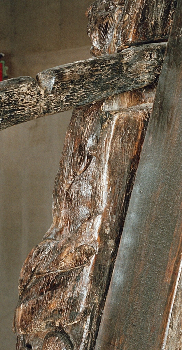 Skulptur i form av en hög, rikt snidad pelare eller stolpe, med kvadratisk genomskärning.

Tre av pelarens sidor är skulpterade medan den fjärde utgör anliggningsyta, försedd med ett antal olikformade urtag. De skulpterade sidorna är horisontellt indelade i tre eller fyra sektioner eller nivåer, avgränsade från varandra av profilerade och snedställda vulster. Nedtill avslutas skulpturen av en ornerad och på mitten insvängd formation.



Ovanför denna nedre avslutning är pelaren utformad som en reslig mansfigur, vänd mot vänster, sedd från betraktaren. Mansfiguren, som täcker tre av skulpturens fyra sidor, är iförd en romersk rustning med en bred fältbindel draperad från höger axel snett över bröstet. På huvudet med det lockiga och utslagna håret sitter en krona av öppen typ, med liljeliknande ornament. Figurens vänstra arm är böjd, medan den högra armen är sträckt utmed sidan. Handen till den högra armen saknas, men har hållit en rikt profilerad sköld, med mycket slitna och vaga konturer av en på bakbenen mot vänster gående grip med stora vingar. Skölden är snidad i ett med figuren, men med kraftiga underskärningar. Vid figurens fötter skymtar ett djur av något slag. Bakom mannens huvud tycks ett snidat textilstycke, kanske del till ett draperi, vara anbragt.



Ovanför den romerska krigaren eller riddaren har pelaren formen av en herm utan armar med långt, krusigt hår och mustaschprytt ansikte. Liksom mansfiguren täcker även hermen tre sidor av pelaren. Huvudet är vridet mot vänster. Överkroppen är bar så när som på ett flikigt plagg som täcker axlar och övre delen av bröstet. Från huvudet och runt midjan är ett tygstycke draperat. Hermens nederdel är formad till en stor maskaron med uppspärrad mun och utsträckt tunga. Maskaronen övergår upptill och utmed sidorna i yppiga bladformationer.



Pelaren avslutas upptill av ett knoppliknande ornament. Ornamentet är ornerat med akantusbladliknande bildningar samt fyra byglar som möts på toppen, där vissa spår antyder att det ursprungligen suttit ett klot eller dylikt, men som fallit bort. Eventuellt kan ornamentet utgöra en huvudbonad till hermen.

Skulpturen är kraftigt sliten.



Text in English: Sculpture in the shape of a richly carved column or pillar.

Three sides of the pillar are carved, while the fourth side comprises the attachment surface and has a number of variously-shaped indentations. The sculptured sides are divided horizontally into three or four sections separated by angled tori.



The lower section of the pillar is carved in the figure of a tall man. The figure is clad in Roman armour and has a field badge (cloak/plaid) draped across his chest at an angle from his right shoulder. On his head he wears an open crown. He has long, curly hair. The right arm is broken off, the left is at his side. The left hand is missing but once held a richly carved shield.

The shield is carved out of the same piece of wood as the figure, but deeply undercut. A carved figure is apparent on the shield. Although the shield is badly worn, it is possible to recognise one of the hind legs of a griffin with large wings. At the man's feet lies an animal of some kind.



Above the Roman warrior or knight, the pillar is carved in the shape of a herm, without arms and with long, curly hair and a moustache. Like the male figure, the herm also covers three sides of the pillar. The head is twisted to the left. The upper part of the body is bare except for a piece of cloth that covers the shoulders and upper chest. The lower part of the herm is carved as a large mask, its mouth open and tongue protruding. The sides and upper parts of the mask are carved as luxuriant leaves.



The upper part of the pillar has a ornament in shape of a knob. The ornament has a ring of acanthus leaves and four spherical loops run vertically from the acanthus leaves and meet at a point at the top. An object, for example a globe appears to have adorned the top of the ornament. This object is missing. The ornament may also be a part of the headgear of the herm.

The sculpture is severely worn.
