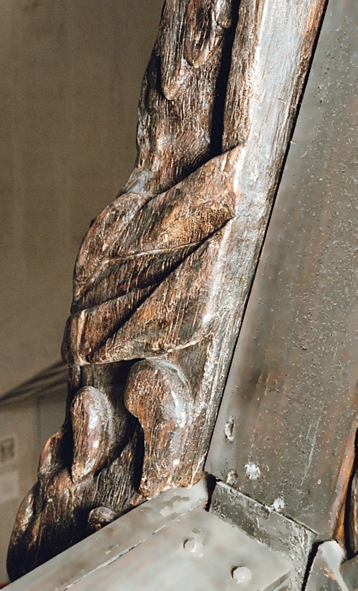 Skulptur i form av en hög, rikt snidad pelare eller stolpe, med kvadratisk genomskärning.

Tre av pelarens sidor är skulpterade medan den fjärde utgör anliggningsyta, försedd med ett antal olikformade urtag. De skulpterade sidorna är horisontellt indelade i tre eller fyra sektioner eller nivåer, avgränsade från varandra av profilerade och snedställda vulster. Nedtill avslutas skulpturen av en ornerad och på mitten insvängd formation.



Ovanför denna nedre avslutning är pelaren utformad som en reslig mansfigur, vänd mot vänster, sedd från betraktaren. Mansfiguren, som täcker tre av skulpturens fyra sidor, är iförd en romersk rustning med en bred fältbindel draperad från höger axel snett över bröstet. På huvudet med det lockiga och utslagna håret sitter en krona av öppen typ, med liljeliknande ornament. Figurens vänstra arm är böjd, medan den högra armen är sträckt utmed sidan. Handen till den högra armen saknas, men har hållit en rikt profilerad sköld, med mycket slitna och vaga konturer av en på bakbenen mot vänster gående grip med stora vingar. Skölden är snidad i ett med figuren, men med kraftiga underskärningar. Vid figurens fötter skymtar ett djur av något slag. Bakom mannens huvud tycks ett snidat textilstycke, kanske del till ett draperi, vara anbragt.



Ovanför den romerska krigaren eller riddaren har pelaren formen av en herm utan armar med långt, krusigt hår och mustaschprytt ansikte. Liksom mansfiguren täcker även hermen tre sidor av pelaren. Huvudet är vridet mot vänster. Överkroppen är bar så när som på ett flikigt plagg som täcker axlar och övre delen av bröstet. Från huvudet och runt midjan är ett tygstycke draperat. Hermens nederdel är formad till en stor maskaron med uppspärrad mun och utsträckt tunga. Maskaronen övergår upptill och utmed sidorna i yppiga bladformationer.



Pelaren avslutas upptill av ett knoppliknande ornament. Ornamentet är ornerat med akantusbladliknande bildningar samt fyra byglar som möts på toppen, där vissa spår antyder att det ursprungligen suttit ett klot eller dylikt, men som fallit bort. Eventuellt kan ornamentet utgöra en huvudbonad till hermen.

Skulpturen är kraftigt sliten.



Text in English: Sculpture in the shape of a richly carved column or pillar.

Three sides of the pillar are carved, while the fourth side comprises the attachment surface and has a number of variously-shaped indentations. The sculptured sides are divided horizontally into three or four sections separated by angled tori.



The lower section of the pillar is carved in the figure of a tall man. The figure is clad in Roman armour and has a field badge (cloak/plaid) draped across his chest at an angle from his right shoulder. On his head he wears an open crown. He has long, curly hair. The right arm is broken off, the left is at his side. The left hand is missing but once held a richly carved shield.

The shield is carved out of the same piece of wood as the figure, but deeply undercut. A carved figure is apparent on the shield. Although the shield is badly worn, it is possible to recognise one of the hind legs of a griffin with large wings. At the man's feet lies an animal of some kind.



Above the Roman warrior or knight, the pillar is carved in the shape of a herm, without arms and with long, curly hair and a moustache. Like the male figure, the herm also covers three sides of the pillar. The head is twisted to the left. The upper part of the body is bare except for a piece of cloth that covers the shoulders and upper chest. The lower part of the herm is carved as a large mask, its mouth open and tongue protruding. The sides and upper parts of the mask are carved as luxuriant leaves.



The upper part of the pillar has a ornament in shape of a knob. The ornament has a ring of acanthus leaves and four spherical loops run vertically from the acanthus leaves and meet at a point at the top. An object, for example a globe appears to have adorned the top of the ornament. This object is missing. The ornament may also be a part of the headgear of the herm.

The sculpture is severely worn.