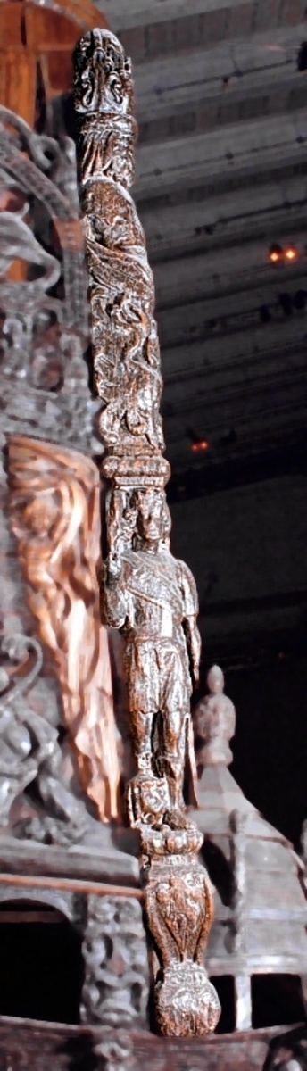 Skulptur i form av en hög, rikt snidad pelare eller stolpe, med kvadratisk genomskärning.

Tre av pelarens sidor är skulpterade medan den fjärde utgör anliggningsyta, försedd med ett antal olikformade urtag. De skulpterade sidorna är horisontellt indelade i tre eller fyra sektioner eller nivåer, avgränsade från varandra av profilerade och snedställda vulster. Nedtill avslutas skulpturen av en ornerad och på mitten insvängd formation.



Ovanför denna nedre avslutning är pelaren utformad som en reslig mansfigur, vänd mot vänster, sedd från betraktaren. Mansfiguren, som täcker tre av skulpturens fyra sidor, är iförd en romersk rustning med en bred fältbindel draperad från höger axel snett över bröstet. På huvudet med det lockiga och utslagna håret sitter en krona av öppen typ, med liljeliknande ornament. Figurens vänstra arm är böjd, medan den högra armen är sträckt utmed sidan. Handen till den högra armen saknas, men har hållit en rikt profilerad sköld, med mycket slitna och vaga konturer av en på bakbenen mot vänster gående grip med stora vingar. Skölden är snidad i ett med figuren, men med kraftiga underskärningar. Vid figurens fötter skymtar ett djur av något slag. Bakom mannens huvud tycks ett snidat textilstycke, kanske del till ett draperi, vara anbragt.



Ovanför den romerska krigaren eller riddaren har pelaren formen av en herm utan armar med långt, krusigt hår och mustaschprytt ansikte. Liksom mansfiguren täcker även hermen tre sidor av pelaren. Huvudet är vridet mot vänster. Överkroppen är bar så när som på ett flikigt plagg som täcker axlar och övre delen av bröstet. Från huvudet och runt midjan är ett tygstycke draperat. Hermens nederdel är formad till en stor maskaron med uppspärrad mun och utsträckt tunga. Maskaronen övergår upptill och utmed sidorna i yppiga bladformationer.



Pelaren avslutas upptill av ett knoppliknande ornament. Ornamentet är ornerat med akantusbladliknande bildningar samt fyra byglar som möts på toppen, där vissa spår antyder att det ursprungligen suttit ett klot eller dylikt, men som fallit bort. Eventuellt kan ornamentet utgöra en huvudbonad till hermen.

Skulpturen är kraftigt sliten.



Text in English: Sculpture in the shape of a richly carved column or pillar.

Three sides of the pillar are carved, while the fourth side comprises the attachment surface and has a number of variously-shaped indentations. The sculptured sides are divided horizontally into three or four sections separated by angled tori.



The lower section of the pillar is carved in the figure of a tall man. The figure is clad in Roman armour and has a field badge (cloak/plaid) draped across his chest at an angle from his right shoulder. On his head he wears an open crown. He has long, curly hair. The right arm is broken off, the left is at his side. The left hand is missing but once held a richly carved shield.

The shield is carved out of the same piece of wood as the figure, but deeply undercut. A carved figure is apparent on the shield. Although the shield is badly worn, it is possible to recognise one of the hind legs of a griffin with large wings. At the man's feet lies an animal of some kind.



Above the Roman warrior or knight, the pillar is carved in the shape of a herm, without arms and with long, curly hair and a moustache. Like the male figure, the herm also covers three sides of the pillar. The head is twisted to the left. The upper part of the body is bare except for a piece of cloth that covers the shoulders and upper chest. The lower part of the herm is carved as a large mask, its mouth open and tongue protruding. The sides and upper parts of the mask are carved as luxuriant leaves.



The upper part of the pillar has a ornament in shape of a knob. The ornament has a ring of acanthus leaves and four spherical loops run vertically from the acanthus leaves and meet at a point at the top. An object, for example a globe appears to have adorned the top of the ornament. This object is missing. The ornament may also be a part of the headgear of the herm.

The sculpture is severely worn.
