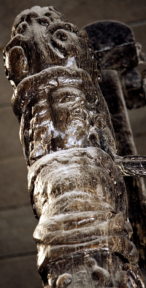 Skulptur i form av en hög, rikt snidad pelare eller stolpe, med kvadratisk genomskärning.

Tre av pelarens sidor är skulpterade medan den fjärde utgör anliggningsyta, försedd med ett antal olikformade urtag. De skulpterade sidorna är horisontellt indelade i tre eller fyra sektioner eller nivåer, avgränsade från varandra av profilerade och snedställda vulster. Nedtill avslutas skulpturen av en ornerad och på mitten insvängd formation.



Ovanför denna nedre avslutning är pelaren utformad som en reslig mansfigur, vänd mot vänster, sedd från betraktaren. Mansfiguren, som täcker tre av skulpturens fyra sidor, är iförd en romersk rustning med en bred fältbindel draperad från höger axel snett över bröstet. På huvudet med det lockiga och utslagna håret sitter en krona av öppen typ, med liljeliknande ornament. Figurens vänstra arm är böjd, medan den högra armen är sträckt utmed sidan. Handen till den högra armen saknas, men har hållit en rikt profilerad sköld, med mycket slitna och vaga konturer av en på bakbenen mot vänster gående grip med stora vingar. Skölden är snidad i ett med figuren, men med kraftiga underskärningar. Vid figurens fötter skymtar ett djur av något slag. Bakom mannens huvud tycks ett snidat textilstycke, kanske del till ett draperi, vara anbragt.



Ovanför den romerska krigaren eller riddaren har pelaren formen av en herm utan armar med långt, krusigt hår och mustaschprytt ansikte. Liksom mansfiguren täcker även hermen tre sidor av pelaren. Huvudet är vridet mot vänster. Överkroppen är bar så när som på ett flikigt plagg som täcker axlar och övre delen av bröstet. Från huvudet och runt midjan är ett tygstycke draperat. Hermens nederdel är formad till en stor maskaron med uppspärrad mun och utsträckt tunga. Maskaronen övergår upptill och utmed sidorna i yppiga bladformationer.



Pelaren avslutas upptill av ett knoppliknande ornament. Ornamentet är ornerat med akantusbladliknande bildningar samt fyra byglar som möts på toppen, där vissa spår antyder att det ursprungligen suttit ett klot eller dylikt, men som fallit bort. Eventuellt kan ornamentet utgöra en huvudbonad till hermen.

Skulpturen är kraftigt sliten.



Text in English: Sculpture in the shape of a richly carved column or pillar.

Three sides of the pillar are carved, while the fourth side comprises the attachment surface and has a number of variously-shaped indentations. The sculptured sides are divided horizontally into three or four sections separated by angled tori.



The lower section of the pillar is carved in the figure of a tall man. The figure is clad in Roman armour and has a field badge (cloak/plaid) draped across his chest at an angle from his right shoulder. On his head he wears an open crown. He has long, curly hair. The right arm is broken off, the left is at his side. The left hand is missing but once held a richly carved shield.

The shield is carved out of the same piece of wood as the figure, but deeply undercut. A carved figure is apparent on the shield. Although the shield is badly worn, it is possible to recognise one of the hind legs of a griffin with large wings. At the man's feet lies an animal of some kind.



Above the Roman warrior or knight, the pillar is carved in the shape of a herm, without arms and with long, curly hair and a moustache. Like the male figure, the herm also covers three sides of the pillar. The head is twisted to the left. The upper part of the body is bare except for a piece of cloth that covers the shoulders and upper chest. The lower part of the herm is carved as a large mask, its mouth open and tongue protruding. The sides and upper parts of the mask are carved as luxuriant leaves.



The upper part of the pillar has a ornament in shape of a knob. The ornament has a ring of acanthus leaves and four spherical loops run vertically from the acanthus leaves and meet at a point at the top. An object, for example a globe appears to have adorned the top of the ornament. This object is missing. The ornament may also be a part of the headgear of the herm.

The sculpture is severely worn.