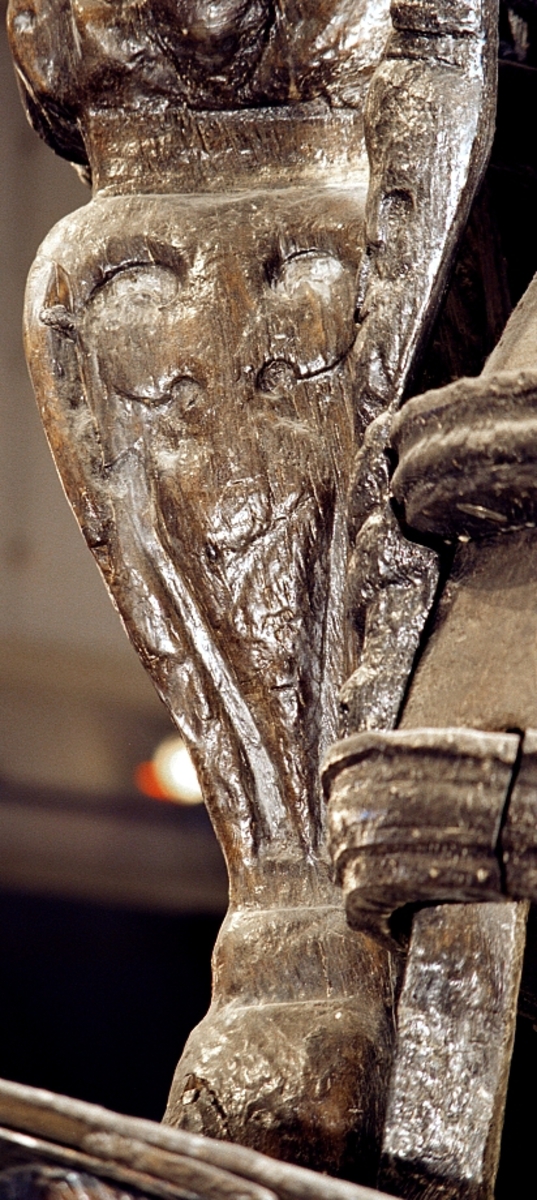 Skulptur i form av en hög, rikt snidad pelare eller stolpe, med kvadratisk genomskärning.

Tre av pelarens sidor är skulpterade medan den fjärde utgör anliggningsyta, försedd med ett antal olikformade urtag. De skulpterade sidorna är horisontellt indelade i tre eller fyra sektioner eller nivåer, avgränsade från varandra av profilerade och snedställda vulster. Nedtill avslutas skulpturen av en ornerad och på mitten insvängd formation.



Ovanför denna nedre avslutning är pelaren utformad som en reslig mansfigur, vänd mot vänster, sedd från betraktaren. Mansfiguren, som täcker tre av skulpturens fyra sidor, är iförd en romersk rustning med en bred fältbindel draperad från höger axel snett över bröstet. På huvudet med det lockiga och utslagna håret sitter en krona av öppen typ, med liljeliknande ornament. Figurens vänstra arm är böjd, medan den högra armen är sträckt utmed sidan. Handen till den högra armen saknas, men har hållit en rikt profilerad sköld, med mycket slitna och vaga konturer av en på bakbenen mot vänster gående grip med stora vingar. Skölden är snidad i ett med figuren, men med kraftiga underskärningar. Vid figurens fötter skymtar ett djur av något slag. Bakom mannens huvud tycks ett snidat textilstycke, kanske del till ett draperi, vara anbragt.



Ovanför den romerska krigaren eller riddaren har pelaren formen av en herm utan armar med långt, krusigt hår och mustaschprytt ansikte. Liksom mansfiguren täcker även hermen tre sidor av pelaren. Huvudet är vridet mot vänster. Överkroppen är bar så när som på ett flikigt plagg som täcker axlar och övre delen av bröstet. Från huvudet och runt midjan är ett tygstycke draperat. Hermens nederdel är formad till en stor maskaron med uppspärrad mun och utsträckt tunga. Maskaronen övergår upptill och utmed sidorna i yppiga bladformationer.



Pelaren avslutas upptill av ett knoppliknande ornament. Ornamentet är ornerat med akantusbladliknande bildningar samt fyra byglar som möts på toppen, där vissa spår antyder att det ursprungligen suttit ett klot eller dylikt, men som fallit bort. Eventuellt kan ornamentet utgöra en huvudbonad till hermen.

Skulpturen är kraftigt sliten.



Text in English: Sculpture in the shape of a richly carved column or pillar.

Three sides of the pillar are carved, while the fourth side comprises the attachment surface and has a number of variously-shaped indentations. The sculptured sides are divided horizontally into three or four sections separated by angled tori.



The lower section of the pillar is carved in the figure of a tall man. The figure is clad in Roman armour and has a field badge (cloak/plaid) draped across his chest at an angle from his right shoulder. On his head he wears an open crown. He has long, curly hair. The right arm is broken off, the left is at his side. The left hand is missing but once held a richly carved shield.

The shield is carved out of the same piece of wood as the figure, but deeply undercut. A carved figure is apparent on the shield. Although the shield is badly worn, it is possible to recognise one of the hind legs of a griffin with large wings. At the man's feet lies an animal of some kind.



Above the Roman warrior or knight, the pillar is carved in the shape of a herm, without arms and with long, curly hair and a moustache. Like the male figure, the herm also covers three sides of the pillar. The head is twisted to the left. The upper part of the body is bare except for a piece of cloth that covers the shoulders and upper chest. The lower part of the herm is carved as a large mask, its mouth open and tongue protruding. The sides and upper parts of the mask are carved as luxuriant leaves.



The upper part of the pillar has a ornament in shape of a knob. The ornament has a ring of acanthus leaves and four spherical loops run vertically from the acanthus leaves and meet at a point at the top. An object, for example a globe appears to have adorned the top of the ornament. This object is missing. The ornament may also be a part of the headgear of the herm.

The sculpture is severely worn.
