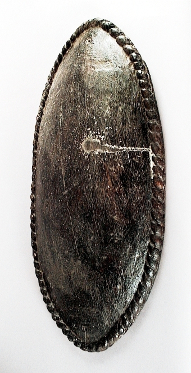 Skulpterad stor, oval och bukig sköld. Skölden pryds av en enkel och tunn kantbård, i form av ett snott band. Framsidan är slät, liksom den urgröpta baksidan. I den ena änden finns ett hål med ett runt märke efter spikhuvud. I den andra änden finns två hål, med fyrkantiga spår av spikhuvuden. Samtliga hål är placerade innanför kantbården.
Skölden är välbevarad, förutom några mindre sprickor.

Text in English: A sculpted large bulging oval shield.
The shield is decorated with a single cord edging, in the form of a slanting band. Front and back are smooth. The back is hollowed out.  At one end of the shield is a hole with the mark from a nail head, at the other end are two holes, with marks of square nail heads. All the holes are situated within the border.
The shield is well preserved, except for a few small cracks.