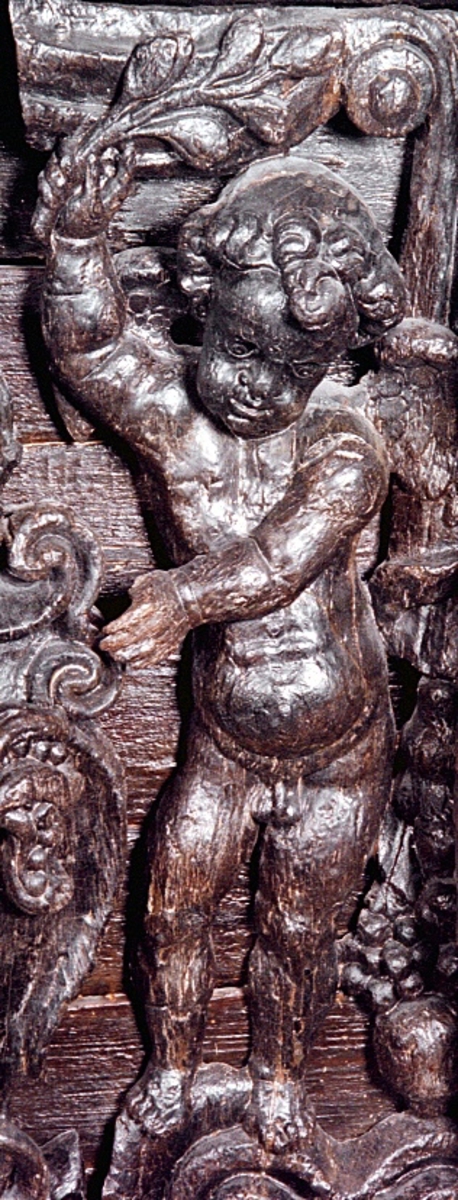 Skulptur av en putto stående på postament med omgivande ornamentik.
Putton har ett frodigt och undersätsigt utseende, med avrundade former. Puttons vänstra hand vilar på en sköld, medan höger hand är uppåtsträckt, hållande en olivkvist.
Ovanför putton sitter ett veckrikt draperi, "upphängt" på en stång med profilerad ände. Där det nedhängande draperiet slutar vidtar tunga fruktfestoner som når ned till postamentet, vilket är dekorerat med symmetriskt ställda rullverksspiraler. Rullverksspiralerna är utformade så att de bildar krumma vädurs- eller gumsehorn till ett i postamentet utsnidat djurhuvud.
Puttons ansikte, vänster arm samt mage är snidade separat. Skulpturens baksida är slät.

Text in English: Sculpture of a putto, standing on a base surrounded by ornamentation.
The exuberant plump cherub has a well-rounded body. Right hand rests on a shield. The left hand holds an olive branch aloft.

Above the putto''s head hangs a heavily-folded drapery. The drapery forms a swag as it hangs from a curtain rod. The prominent ends of the curtain rod are carved in relief. Heavy festoons of fruit flow from the drapery ends to the feet of the putto. The symmetrical scrollwork on the base takes on the appearance of ram''s horns when combined with the imprecise representations of animal heads.
Face, left arm and belly are carved separately. Reverse side is smooth.