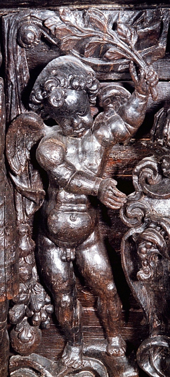 Skulpterad putto figur stående på postament med omgivande ornamentik.
Putton har ett frodigt och undersätsigt utseende, med avrundade former. Puttons vänstra hand vilar på en sköld, medan höger hand är uppåtsträckt, hållande en olivkvist.
Ovanför putton sitter ett veckrikt draperi, "upphängt" på en stång med profilerad ände. Där det nedhängande draperiet slutar vidtar tunga fruktfestoner. Festonerna når ned till postamentet, som är dekorerat med symmetriskt ställda rullverksspiraler. Rullverksspiralerna är utformade så att de bildar krumma vädurs- eller gumsehorn till ett i postamentet utsnidat djurhuvud. Skulpturens baksida är slät.
Puttons ansikte, vänster arm samt mage är snidade separat.

Text in English: Sculpture of a putto, standing on a base surrounded by ornamentation.
The exuberant, plump putto has a rounded body. The putto''s left hand rests on a shield. The right hand is holding an olive branch aloft. Above the putto''s head hangs a heavily-folded drapery.
The drapery forms a swag as it hangs from a curtain rod. The prominent ends of the curtain rod are carved in relief. Heavy festoons of fruit flow from the drapery ends to the feet of the putto. The symmetrical scrollwork on the base takes on the appearance of ram''s horns when combined with the imprecise representations of animal heads. The sculpture''s reverse side is smooth.
Face, right arm and belly are carved separately.