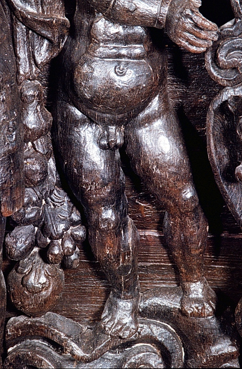 Skulpterad putto figur stående på postament med omgivande ornamentik.
Putton har ett frodigt och undersätsigt utseende, med avrundade former. Puttons vänstra hand vilar på en sköld, medan höger hand är uppåtsträckt, hållande en olivkvist.
Ovanför putton sitter ett veckrikt draperi, "upphängt" på en stång med profilerad ände. Där det nedhängande draperiet slutar vidtar tunga fruktfestoner. Festonerna når ned till postamentet, som är dekorerat med symmetriskt ställda rullverksspiraler. Rullverksspiralerna är utformade så att de bildar krumma vädurs- eller gumsehorn till ett i postamentet utsnidat djurhuvud. Skulpturens baksida är slät.
Puttons ansikte, vänster arm samt mage är snidade separat.

Text in English: Sculpture of a putto, standing on a base surrounded by ornamentation.
The exuberant, plump putto has a rounded body. The putto''s left hand rests on a shield. The right hand is holding an olive branch aloft. Above the putto''s head hangs a heavily-folded drapery.
The drapery forms a swag as it hangs from a curtain rod. The prominent ends of the curtain rod are carved in relief. Heavy festoons of fruit flow from the drapery ends to the feet of the putto. The symmetrical scrollwork on the base takes on the appearance of ram''s horns when combined with the imprecise representations of animal heads. The sculpture''s reverse side is smooth.
Face, right arm and belly are carved separately.