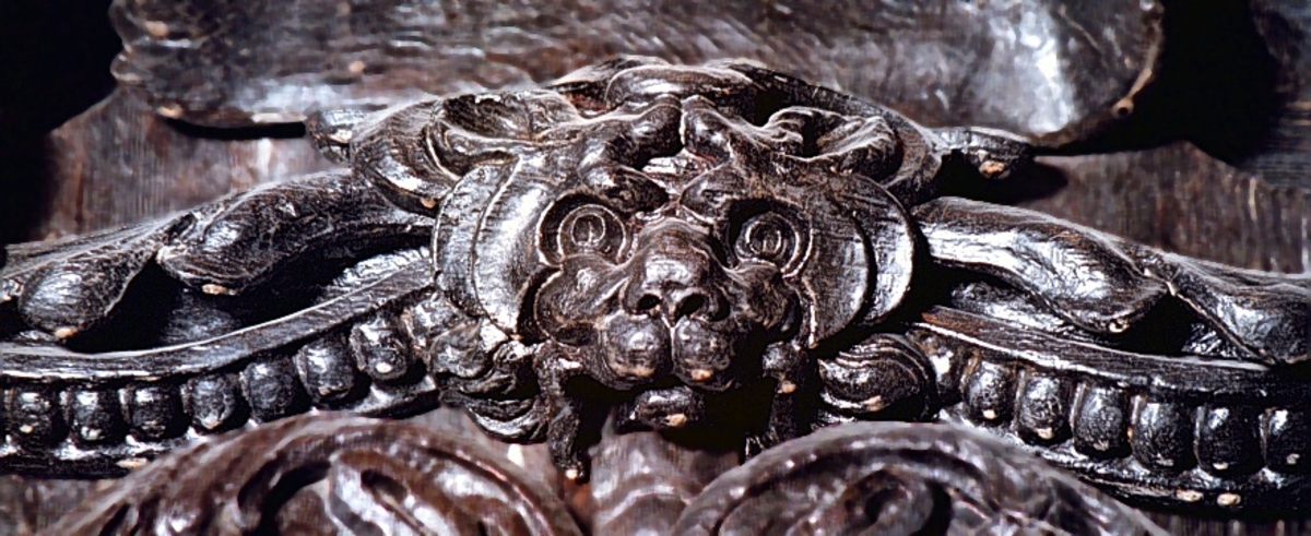 Broskornament. Ornamentet är utformat som ett kattdjursliknande huvud från vilket utgår "armar", dekorerade med blad och broskvoluter. Ändpartierna är förtjockade med elipsformade broskvoluter, blad och nedhängande flikar. Baksidan är slät.
Ornamentet är brutet på baksidans mittparti.

Text in English: Earshell ornament consisting of a cat-like head, from which extend "arms" decorated with ellipse-shaped pearlwork scroll leaves and downward-hanging leaf-blades with thickened ends. The reverse side is smooth.
The ornament is broken in the middle on the reverse side.