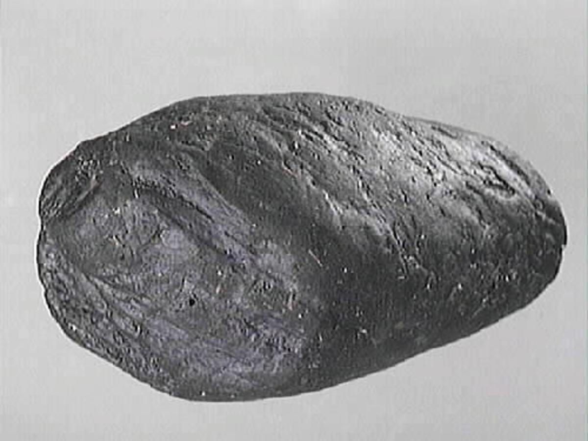 Del av arm till en skulpterad krigare. Oval form. Ovansidan är rundad. Baksidan är troligtvis bruten. Ena kortänden är bruten.
Skulpturdelen är mycket sliten.

Text in English: Part of sculpted arm of a warrior. Oval shaped. Slightly more rounded on one side, one end is broken. Much worn.