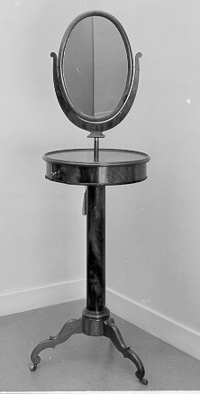 Spegelbord, rakbord, runt med oval spegel i en spegelhållare
i form av en lyra. Spegelns mått; Bredd 300 mm, längd 430 mm. Bordet
vilar på en pelarfot, som i sin tur står på tre fötter, två
halvcirkelformade lådor med lås. Vid vridning av nyckeln åt ett håll,
sprätter den ena lådan ut och vid vridning åt motsatt håll den
andralådan. Bordet har tillhört Postverkets Generaldirektör Adolf
Wilhelm Roos.
