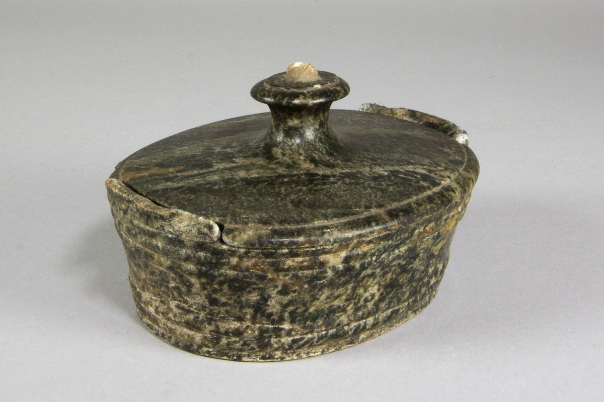 Smörask, med lock, av sten. Oval form med höga, svagt utåtstående, sidor och med två öron på kortsidorna. Med profilerade bårder. Locket med oval form med profilerad kant och med utnsitt för att passa in med askens öron. Med en balusterformad oval knopp.