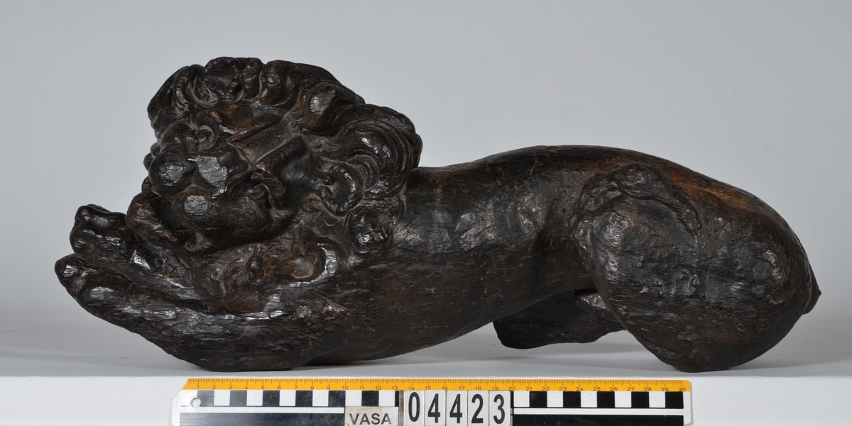 Skulptur av ett litet, liggande lejon med utsträckta framben och hopdragna bakben.
Lejonet är riktat mot vänster, sett från betraktaren. Det snett hållna huvudet återges i det närmaste i en face. Större delen av svansen saknas. Den befintliga delen av svansen avtecknar sig mot lejonets vänstra lår.

Lejonet är endast delvist rundskulpterat. På baksidan, i höjd med huvudet och höger framben, återfinns ett stort, snett urtag som bildar en slät anliggningsyta. Skulpturens framparti är således endast till hälften så tjockt som dess bakparti.
Bakkroppen är kraftigt sliten och skadad. Vänster bakben saknas, liksom större delen av svansen.

Text in English: Sculpture of a small reclining lion with its forelegs extended and its  hind legs tucked beneath its body.
Facing to the observer''s left, the head, held crookedly, is nearly en face. A large part of the tail is missing, and the remainder curves towards the left hind leg.

The lion is only partly three-dimensional. On the rear at the head height and at the right foreleg a large oblique hole forms a flat area for attachement, thus the forepart of the sculpture is thus only half as thick as its rear.
The rear of the body is badly worn and damaged. The left hind leg has been broken off.