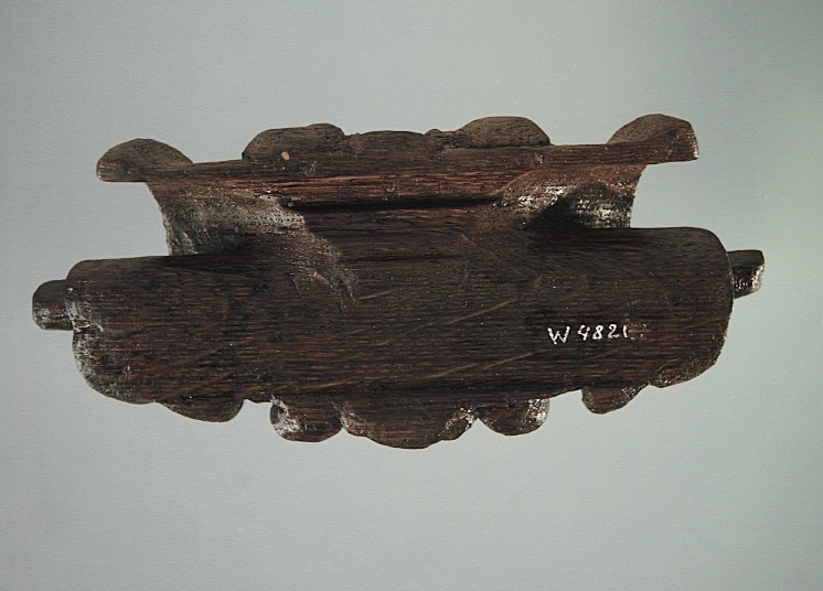 Rullverksornament.
Skulpterat litet, spänneliknande ornament, dekorerat i broskverksstil.
Det långsmala ornamentet är format som ett stycke kraftigt upprullad bit näver eller läder, med konkav bak- eller undersida. På baksidan är ornamentet profilerat för en list. Broskdekoren på ovansidan är formad som en maskaron.
Ornamentet är mycket välbevarat.

Text in English: A small, buckle-like ornament, decorated in a style similar to earshell work.
The long narrow ornament is formed as a piece of tightly rolled bark or leather, with a concave back or underside so as to match a batten or the like. The reverse side is profiled to fit a moulding. The earshell work on the upper side is shaped as a mask.
The ornament is well preserved.