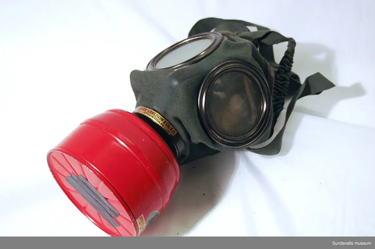 Gasmask/skyddsmask för ansiktet med filter mot föroreningar. Modellnamn 'Degea'. Förvaras i svart cylinderformad plåtbehållare med axelrem i brunt läder. I behållarens lock finns ett fack i vilket två sk. klarskivor förvaras i pappersomslag.  Masken är tillverkad av grönt bomullstyg i kraftig kvalitet, med runda metallinramade glas vid ögonen och en röd filterbehållare i metall. Invändigt är masken fodrad med sämskskinn. Masken spänns fast på huvudet med hjälp av resårband och reglerbara spännremmar.