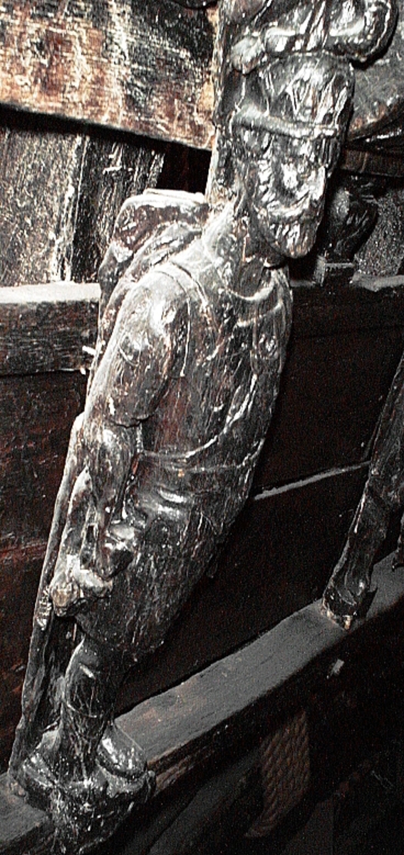 Skulptur föreställande en mansfigur med ett långsmalt och skäggigt ansikte, under en hög, slät hjälm med stora plymer. Runt hjälmen är en stålkrona trädd. Bakom huvudet skymtar ett snäckskal. Figuren är iförd en rikt ornerad rustning av thoraxmodell. Utefter figurens rygg hänger en mantel, som också draperar den vänstra axeln.

Hela figuren är frontalt framställd. Höger arm hålls lätt böjd utefter sidan, med handen fattande om ett svärdsfäste. Vänster arm är något böjd rakt ut från sidan, och handen verkar fatta tag om manteln. Kroppstyngden är jämnt fördelad på båda benen.

Figuren står på en sockel, vilken pryds av en bred och grimaserande maskaron. Sockeln är även försedd med en banderoll som tycks helt slät och utan namninskription.

Upptill är bakstycket framdraget i en kraftig "huvudplatta". Skulpturens baksida uppvisar tre snedställda, olika djupt liggande fält. Fälten, som utgör anliggningsytor, ger skulpturen en trappstegsformad profil.
Huvudplattan är delvis skadad. Svärdets klinga saknas. I övrigt är skulpturen relativt välbevarad.

Text in English: A sculpture representing a male figure with a long and narrow face that is bearded and has a moustache. On the head there is a plumed helmet on which a crown with points has been placed and behind the head a large sea shell. The figure is wearing richly decorated thorax type armour. The mantle is hanging down the back and draped over the left shoulder.

The figure faces towards the front. The right arm is slightly bent and held at the side and the hand holds a sword hilt. The left arm is slightly bent and held out from the side and the left hand seems to hold the mantle. The weight of the body is carried on both legs.

The figure stands on a plinth with a broad and grimacing mask and also has a label which is quite smooth and without any inscription.

The upper part of the back extends obliquely upwards in a powerful "head board". The sculpture''s back has three obliquely placed surfaces of different depths that are areas of attachment. The surfaces make the sculpture look like a step-ladder in profile.
The board behind the head is partly damaged and the blade of the sword is missing but otherwise, the sculpture is rather well preserved.