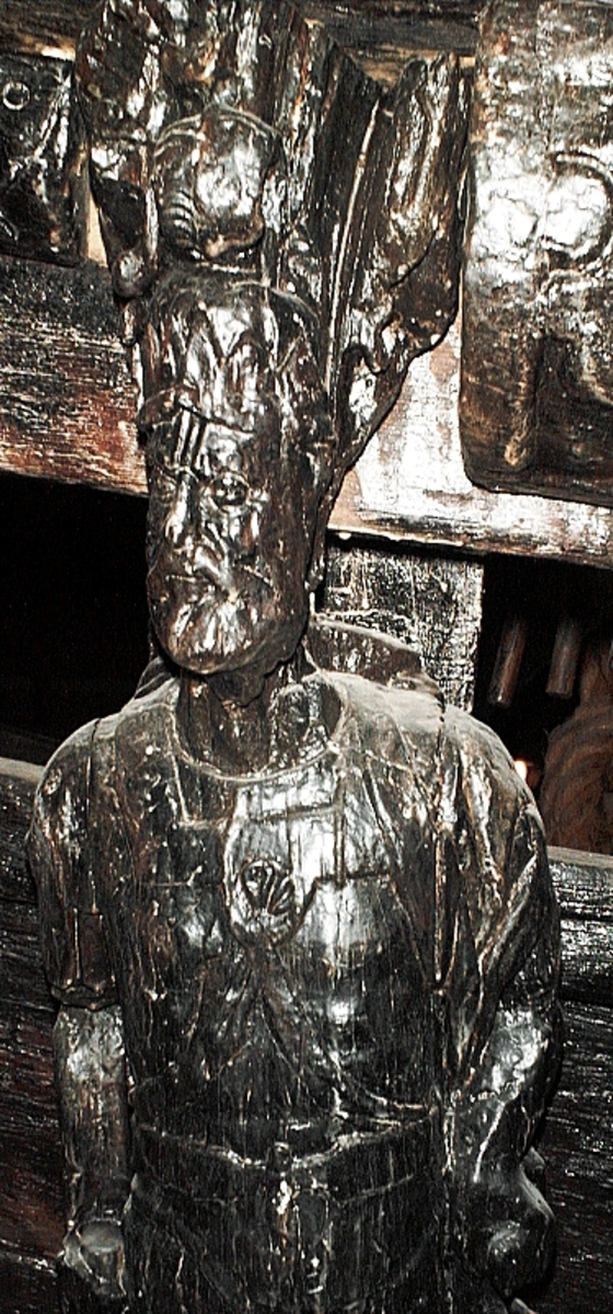 Skulptur föreställande en mansfigur med ett långsmalt och skäggigt ansikte, under en hög, slät hjälm med stora plymer. Runt hjälmen är en stålkrona trädd. Bakom huvudet skymtar ett snäckskal. Figuren är iförd en rikt ornerad rustning av thoraxmodell. Utefter figurens rygg hänger en mantel, som också draperar den vänstra axeln.

Hela figuren är frontalt framställd. Höger arm hålls lätt böjd utefter sidan, med handen fattande om ett svärdsfäste. Vänster arm är något böjd rakt ut från sidan, och handen verkar fatta tag om manteln. Kroppstyngden är jämnt fördelad på båda benen.

Figuren står på en sockel, vilken pryds av en bred och grimaserande maskaron. Sockeln är även försedd med en banderoll som tycks helt slät och utan namninskription.

Upptill är bakstycket framdraget i en kraftig "huvudplatta". Skulpturens baksida uppvisar tre snedställda, olika djupt liggande fält. Fälten, som utgör anliggningsytor, ger skulpturen en trappstegsformad profil.
Huvudplattan är delvis skadad. Svärdets klinga saknas. I övrigt är skulpturen relativt välbevarad.

Text in English: A sculpture representing a male figure with a long and narrow face that is bearded and has a moustache. On the head there is a plumed helmet on which a crown with points has been placed and behind the head a large sea shell. The figure is wearing richly decorated thorax type armour. The mantle is hanging down the back and draped over the left shoulder.

The figure faces towards the front. The right arm is slightly bent and held at the side and the hand holds a sword hilt. The left arm is slightly bent and held out from the side and the left hand seems to hold the mantle. The weight of the body is carried on both legs.

The figure stands on a plinth with a broad and grimacing mask and also has a label which is quite smooth and without any inscription.

The upper part of the back extends obliquely upwards in a powerful "head board". The sculpture''s back has three obliquely placed surfaces of different depths that are areas of attachment. The surfaces make the sculpture look like a step-ladder in profile.
The board behind the head is partly damaged and the blade of the sword is missing but otherwise, the sculpture is rather well preserved.
