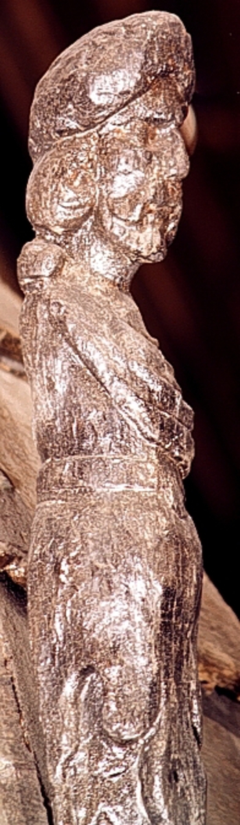 Skulpterad triton med turbanliknande lagerkrans på huvudet.

Huvudet har långt, ringlande hår samt stora och nedhängande mustascher. Snett över bröstet är en klädnad draperad. Klädnaden är fästad i en knut på höger axel. Kring livet sitter ett band med en oval.



Kroppens övergång till de dubbla, snodda fiskstjärtarna döljs av ett stort rundflikigt akantusblad. Huvudet är till största delen framställd som rundskulptur, dock något avplanad. Från nackhöjd och halvvägs ned på partiet med fiskstjärtarna är baksidan avplanad och slät - delvis något skålad - varefter det sitter ett snett urtag. Fotändan är vinkelformigt skålad vilket ger den en V-format tvärsektion, nedsänkningen är dock inte symmetriskt placerad. Nedtill böjer fotänden av inåt.

Skulpturen är mycket välbevarad.



Text in English: Sculpted Triton with a turban-like laurel wreath on its head.

The figure has a large pendulous moustache and long curly hair. There is a garment draped diagonally across the chest and fastened in a knot on the right shoulder. There is a girdle or belt round the waist with an oval centrepiece.



The transition between the body and the double fish tails is concealed by a large rounded acanthus leaf. The majority of the head is carved as a three-dimensional sculpture. The rear side surface has been planed smoothly.  The foot end has been hollowed out at an angle, giving it a V-shaped cross section, the hollowed-out section is not, however, symmetrically placed. The foot end curves inwards.

The sculpture is very well preserved.